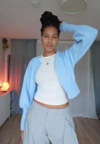 Hellblauer Strickcardigan mit langen, zulaufenden Ärmeln, Knopfverschluss, getragen über einem weißen, figurbetonten Oberteil und grauen, faltengestuften Hosen.