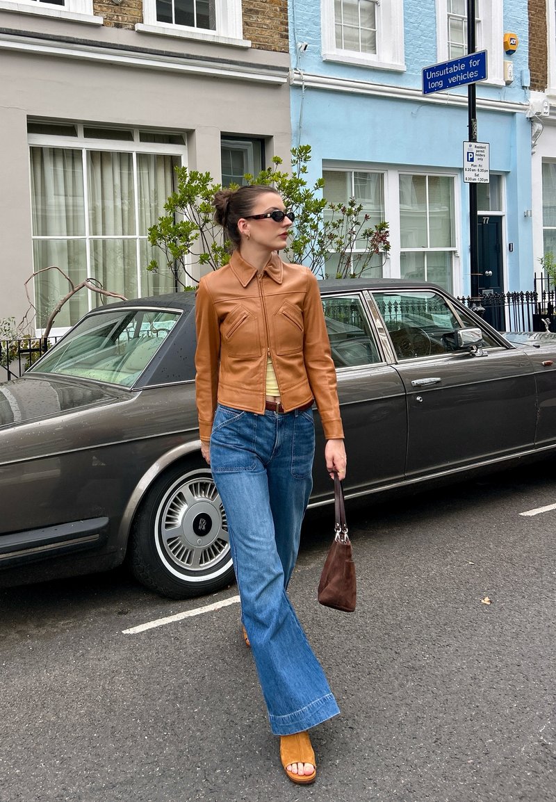 Woman in brown leather jacket and blue jeans walking past a vintage dark grey car on a city street with colourful buildings in the background.