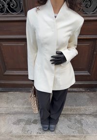 White tailored blazer with a fitted design, long sleeves, and stylish seams, paired with black trousers and leather gloves. Brown patterned handbag.