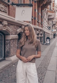 Brun kortärmad topp och vita vida byxor, stående på en kullerstensgata med tegelbyggnader och ett hängande skylt i bakgrunden.