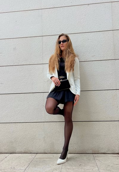 Un vestido negro con una falda acampanada, una chaqueta blanca, un cinturón negro con una hebilla dorada, medias negras y tacones blancos, frente a una pared gris texturizada.