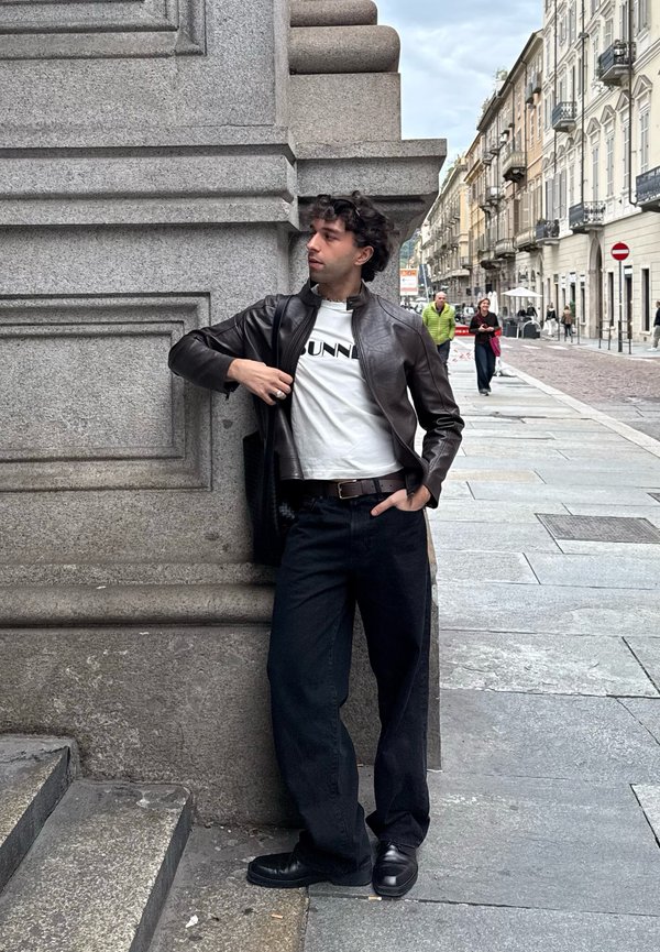 Giacca di pelle marrone sopra una maglietta bianca con testo nero, pantaloni neri e scarpe nere, posizionata contro una colonna architettonica in pietra.