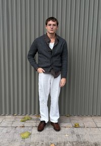 Ung mand med kort hår og briller står foran en grå væg med lodrette paneler, iført en mørk cardigan, brun skjorte, hvide bukser og brune sko.