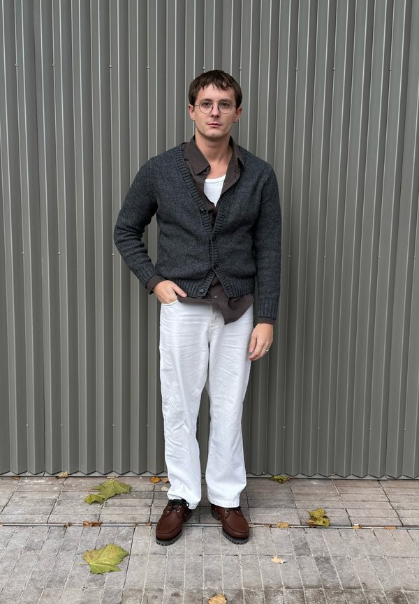 Junger Mann mit kurzen Haaren und Brille steht vor einer grauen Wand mit vertikalen Paneelen, trägt eine dunkle Strickjacke, ein braunes Hemd, weiße Hose und braune Schuhe.