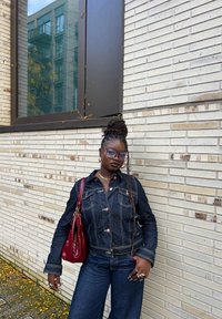 Jeans jakna z zlatimi šivi in gumbi, v kombinaciji s širokimi temno modrimi kavbojkami. Drži svetlo rdečo torbico. Ozadje je nevtralna opečnata stena.