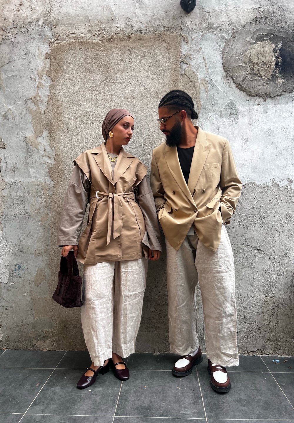 Due persone che si fronteggiano. Una indossa un cappotto beige con cintura e pantaloni di lino; l'altra un blazer beige e pantaloni bianchi a righe.