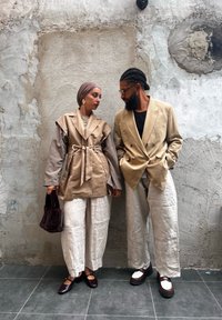 Due persone che si fronteggiano. Una indossa un cappotto beige con cintura e pantaloni di lino; l'altra un blazer beige e pantaloni bianchi a righe.