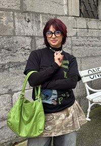 Zelena kožna torba mekog teksture, zaobljenog oblika i s dva ručka. Crna majica s grafičkim printom i slojevita suknja. Pozadina od kamenog zida.