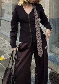 Brūna blūze ar pogu aizdari un dziļu V-veida izgriezumu, kombin�ēta ar vaļīgām brūnām satīna biksēm un rakstainu brūnu kaklasaiti. Iekļauta melna plecu soma.