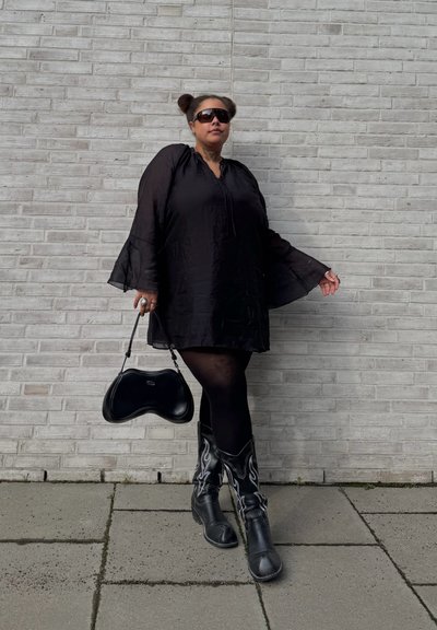 Persona con vestido negro, medias negras, botas negras con estampado, gafas de sol y bolso negro, de pie frente a una pared de ladrillos de color gris claro.