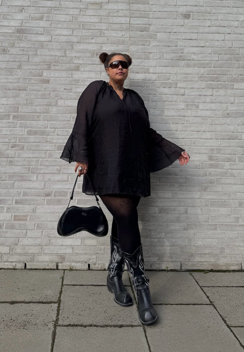 Osoba oblečená v čiernych šatách, čiernych pančuchách, so vzorovanými čiernymi čižmami, slnečnými okuliarmi a držiaca čiernu kabelku, stojaca pri svetlosivej tehlovej stene.
