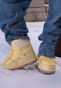 Bottes Moon jaunes avec lacets portées par-dessus un jean bleu, debout sur un sol enneigé près de marches en pierre.