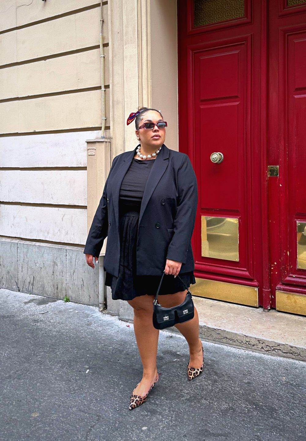 Μαύρο σακάκι πάνω από μαύρη φόρεμα, κολιέ από μαργαριτάρια, μαύρη τσάντα και φλατ παπούτσια με λεοπάρ σχέδιο. �Λαμπερές κόκκινες πόρτες στο υπόβαθρο. Λείο πέτρινο πλακόστρωτο.