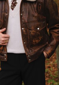Giacca di pelle marrone con colletto in pelliccia, chiusura con zip e bottoni a scatto. Abbinata a un maglione bianco e pantaloni scuri. Superficie texturizzata visibile.