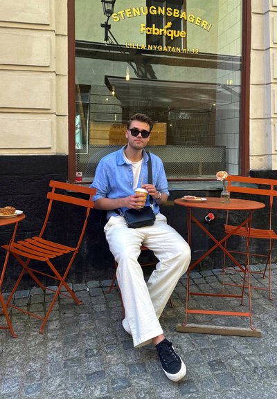 Una camisa de manga corta azul claro contrasta con pantalones anchos blancos. La escena presenta sillas de metal naranjas, un bolso negro y pasteles sobre las mesas.