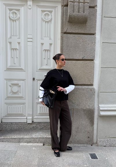 Mujer con top en capas blanco y negro y pantalones oscuros, de pie junto a una puerta blanca ornamentada y una pared de piedra, sosteniendo un bolso negro y llevando gafas de sol.
