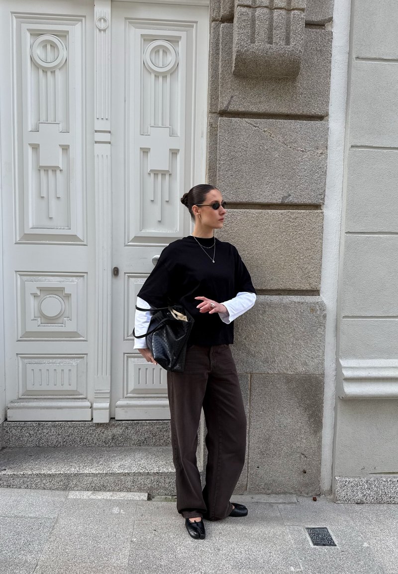 Femme en haut superposé noir et blanc et pantalon foncé, debout près d'une porte blanche ornée et d'un mur en pierre, tenant un sac à main noir et portant des lunettes de soleil.
