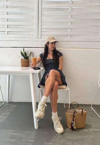 Vestido negro con flores, gorra beige, bebiendo un batido. Mesa blanca, planta en maceta, bolsa de trenzado con borlas, suelo gris, luz natural que entra a través de lamas.
