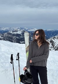 Femme portant des lunettes de soleil et un pull gris tient un casque de ski à côté de skis et bâtons sur une montagne enneigée avec des sommets lointains en arrière-plan.