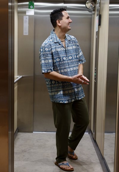 Camisa azul de manga corta con estampado, pantalones verdes y chanclas marrones, de pie en un ascensor con interiores metálicos y beige.