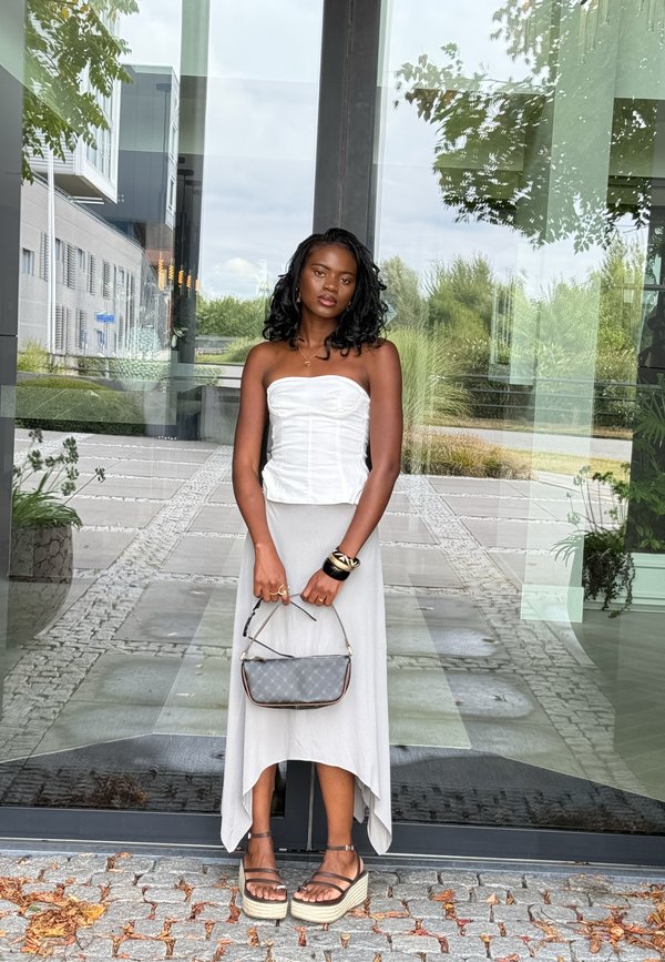 Een vrouw staat voor een glazen ingang. Ze draagt een witte, strapless top en een grijze rok, en houdt een grijze handtas vast. Haar sandalen hebben een plateauzool.