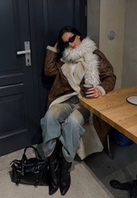 Ruda avies kailio striukė su balta kailio apykakle, mėlyni plyšę džinsai, juodi smailūs batai ir juoda rankinė. Sėdi prie medinio stalo.