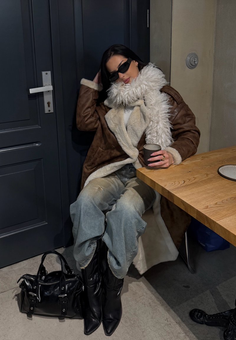 Manteau en shearling marron avec un col en fourrure blanche, jean bleu déchiré, bottes noires pointues et sac à main noir. Assise à une table en bois.