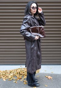 Brauner Leder-Trenchcoat mit Knopfdetails, kombiniert mit einer dunklen Handtasche und schwarzen weiten Hosen. Das Model trägt runde Sonnenbrillen.