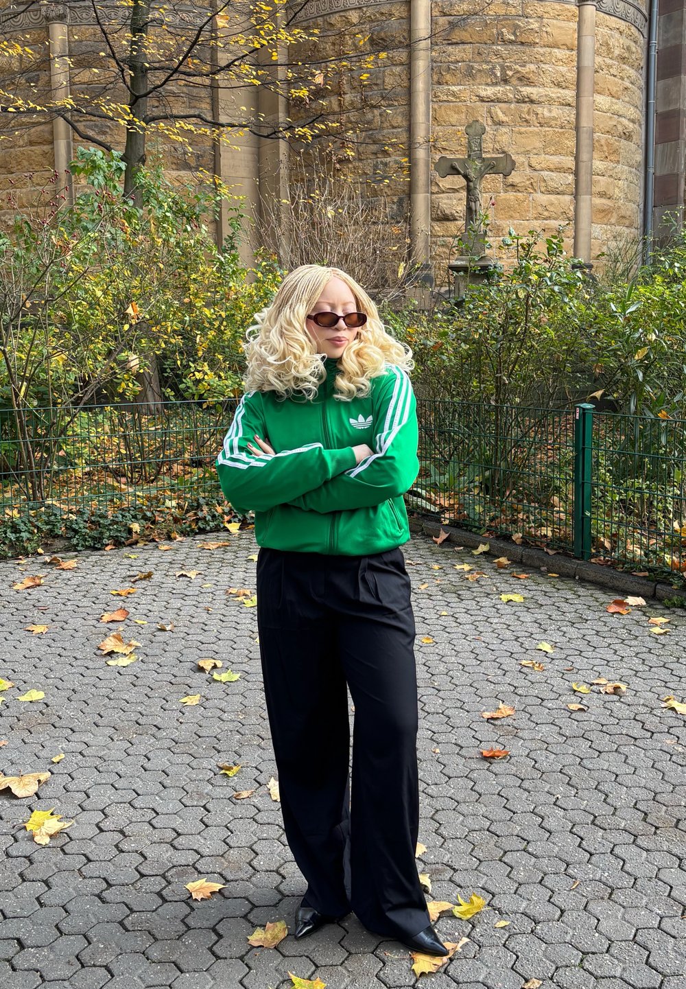 Casaco verde com fecho éclair e riscas brancas, combinado com calças pretas. O casaco apresenta o logótipo da Adidas. Mulher com óculos de sol e cabelo loiro ondulado.