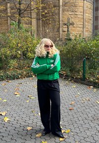 Casaco verde com fecho éclair e riscas brancas, combinado com calças pretas. O casaco apresenta o logótipo da Adidas. Mulher com óculos de sol e cabelo loiro ondulado.