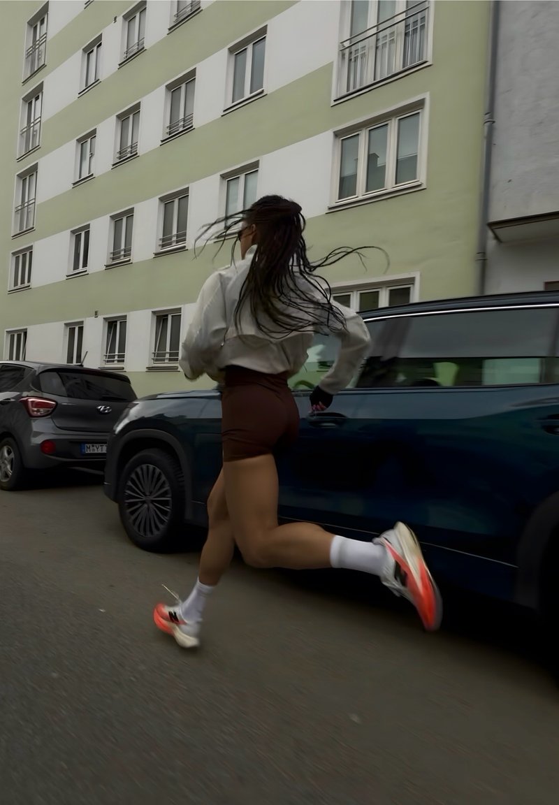 Γυναίκα με μακριά μαλλιά τρέχει πέρα από σταθμευμένα αυτοκίνητα σε έναν αστικό δρόμο δίπλα σε μια πολυώροφη κατοικία.