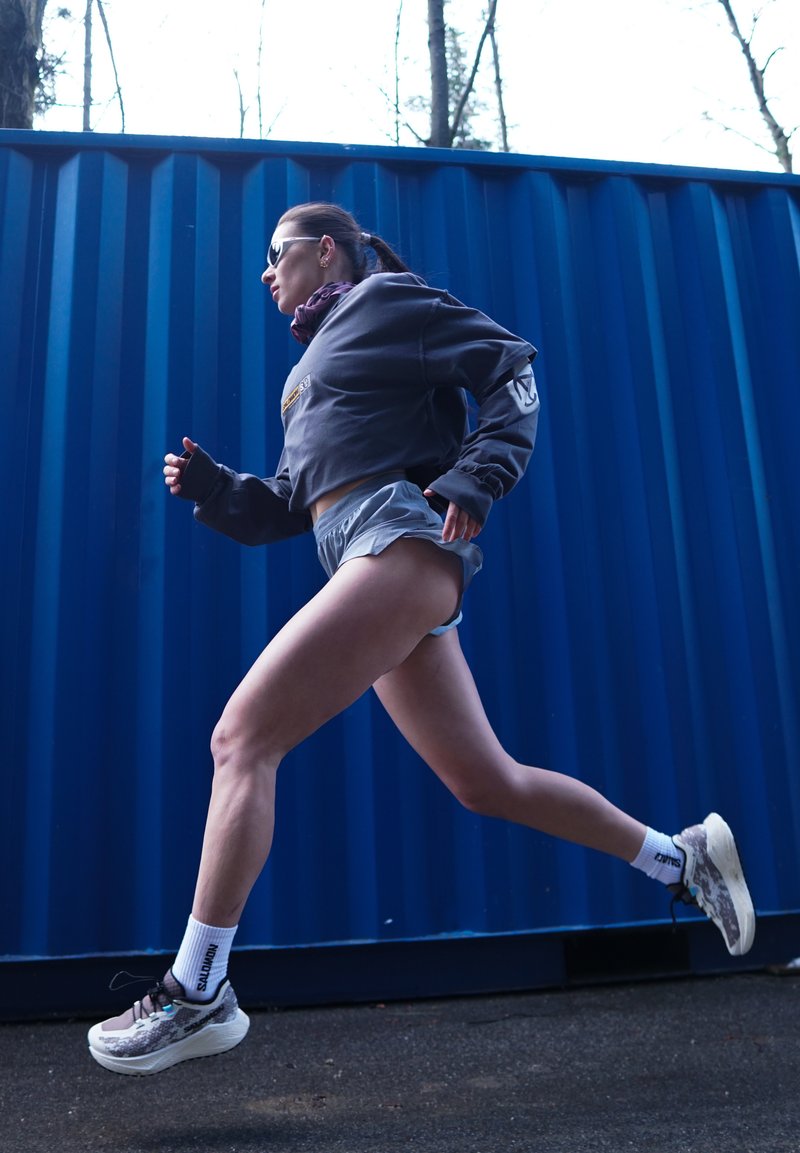Woman wearing sunglasses, grey hoodie, and shorts running past a blue container outdoors on a paved surface.