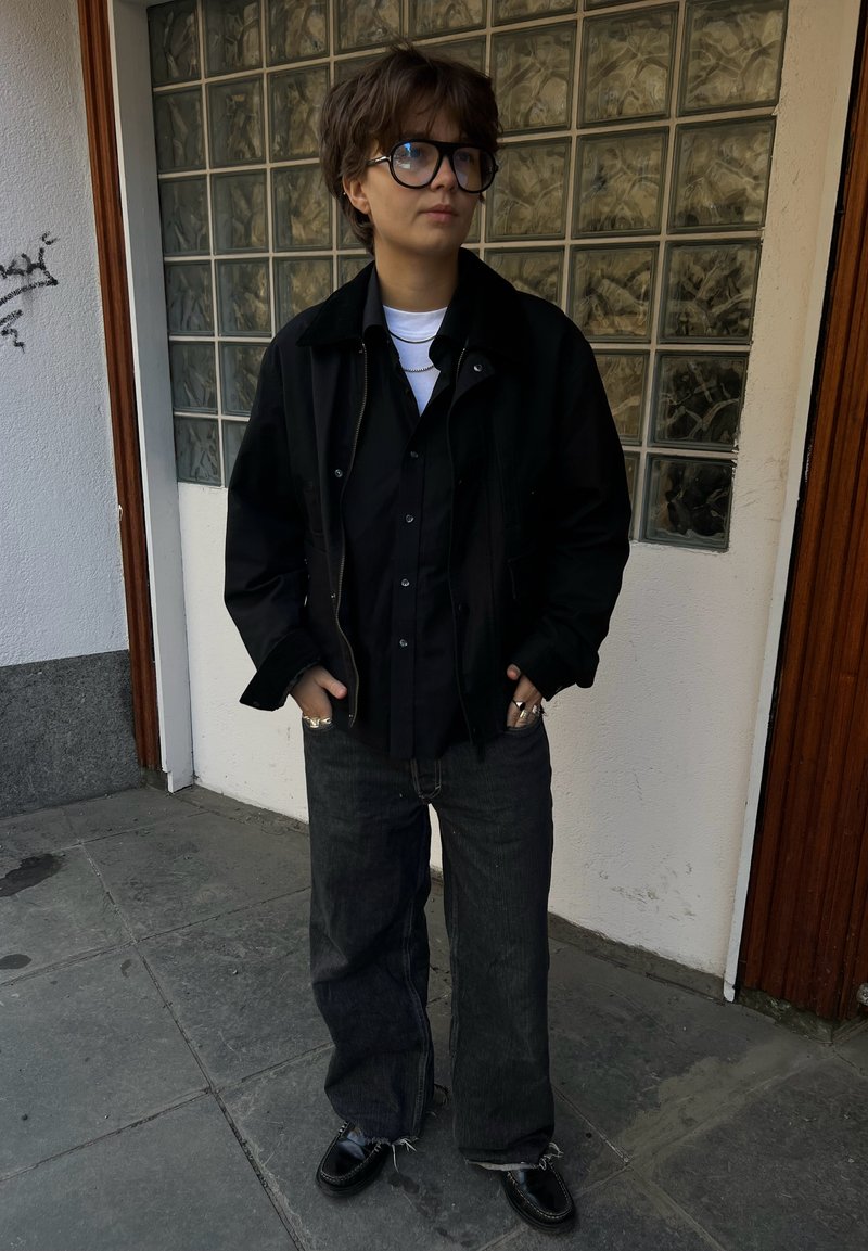 Personne aux cheveux courts et grandes lunettes portant plusieurs vestes noires superposées, un jean foncé et des chaussures noires, debout sur un trottoir carrelé près d’un mur.