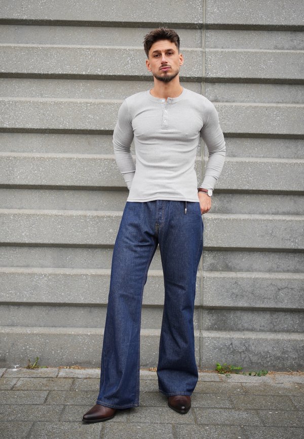 Un homme pose devant un mur en pierre gris. Il porte une chemise gris clair à manches longues, un jean bleu à jambes larges et des chaussures brunes.
