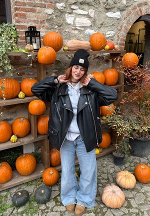 Mujer con gorro negro y chaqueta de cuero sonríe entre calabazas naranjas en estantes de madera afuera de un edificio de piedra.