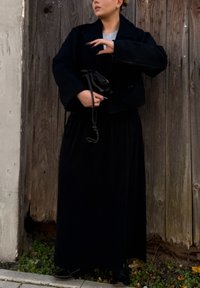 Zwarte cropped jas over een grijze top, gecombineerd met een lange zwarte rok. Een kleine zwarte tas wordt in één hand vastgehouden, met een houten achtergrond.