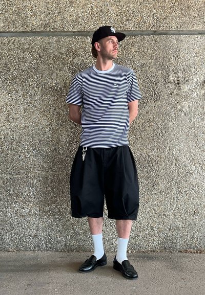 Pantalones cortos negros y holgados, camiseta a rayas en blanco y azul marino, gorra negra, calcetines blancos, zapatos negros, con un detalle de llavero en los pantalones cortos.