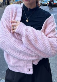 Ljusrosa stickad cardigan med en texturerad, ribbad design. Har vida ärmar och en knappstängning vid fållen. Bärs över en svart topp.