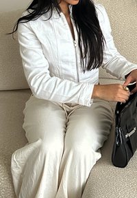 Weiße Reißverschlussjacke und cremefarbene Hose. Glatter Stoff und lockere Passform. Schwarze Handtasche mit silberfarbenen Metallakzenten an der Seite.