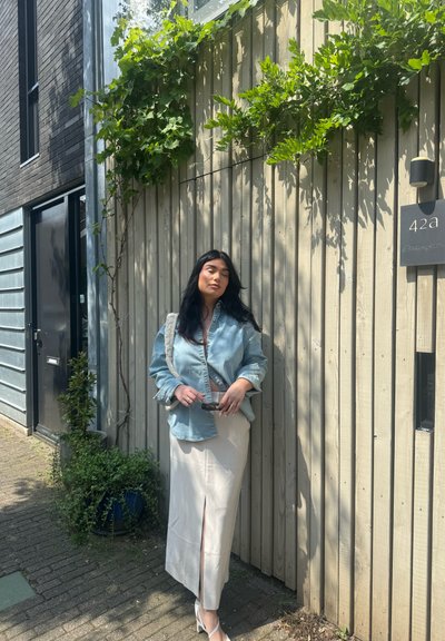 Camisa de mezclilla, azul claro, corte oversize. Falda midi de satén crema con abertura frontal. Tacones blancos. Fondo de valla de madera con follaje verde.
