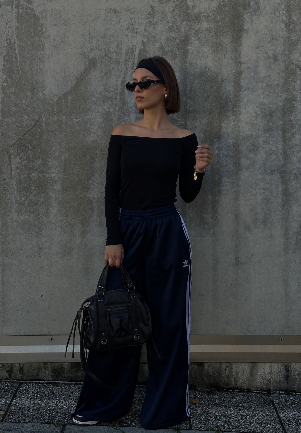Femeie îmbrăcată într-un top negru fără umeri și pantaloni sport bleumarin Adidas stă sprijinită de un perete gri de beton, ținând o geantă neagră de mână.