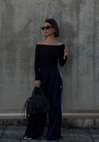 Femeie îmbrăcată într-un top negru fără umeri și pantaloni sport bleumarin Adidas stă sprijinită de un perete gri de beton, ținând o geantă neagră de mână.