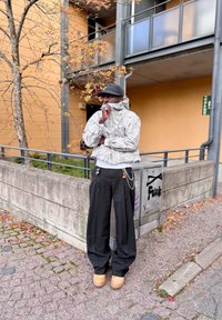 Šedá prešívaná bunda s vzorom, čierne nohavice s širokými nohami, hnedé topánky a doplnky zahŕňajúce reťaze a farebné prívesky. Mestské vonkajšie prostredie.