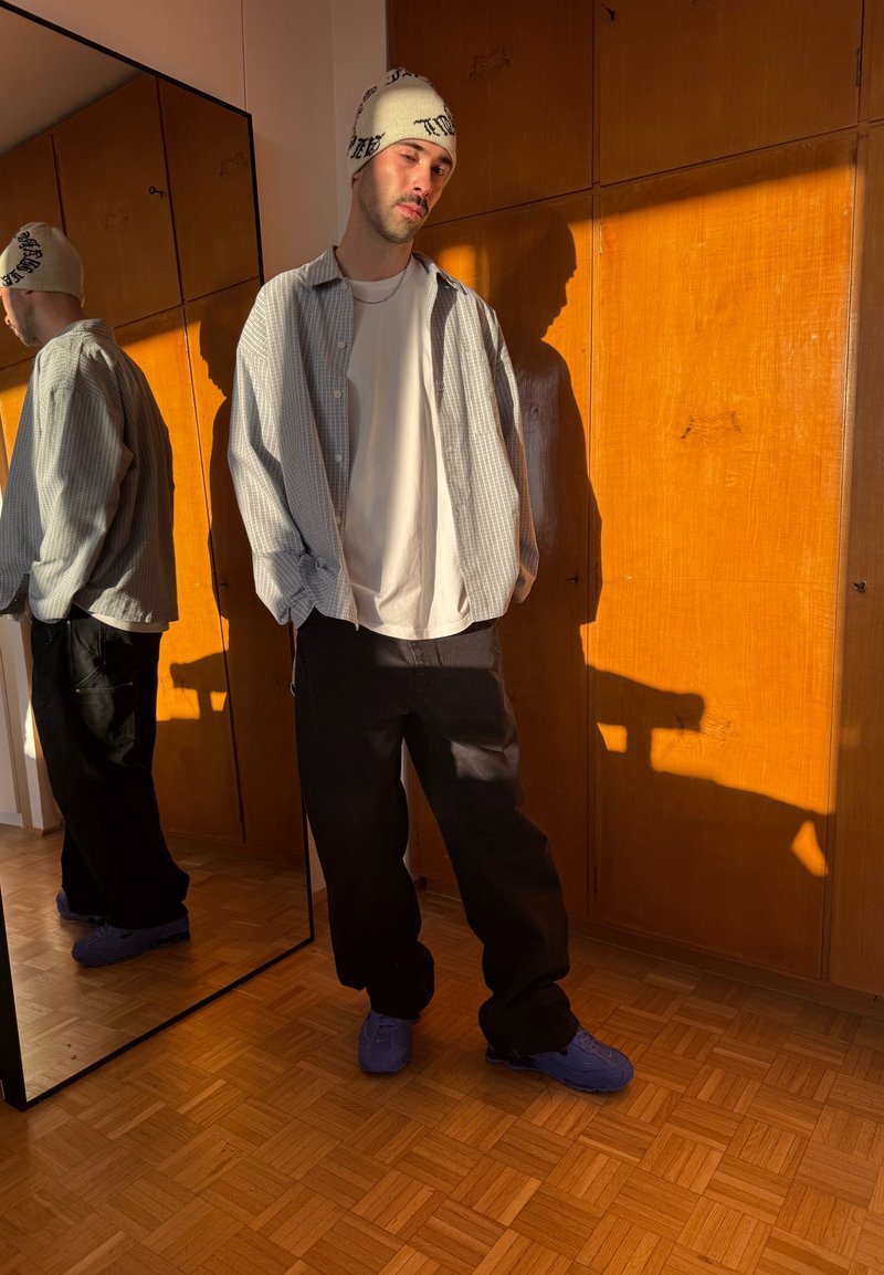 Joven con ropa casual y gorro de lana, de pie con las manos en los bolsillos, apoyado contra una pared de madera, con su reflejo en un espejo alto cercano.