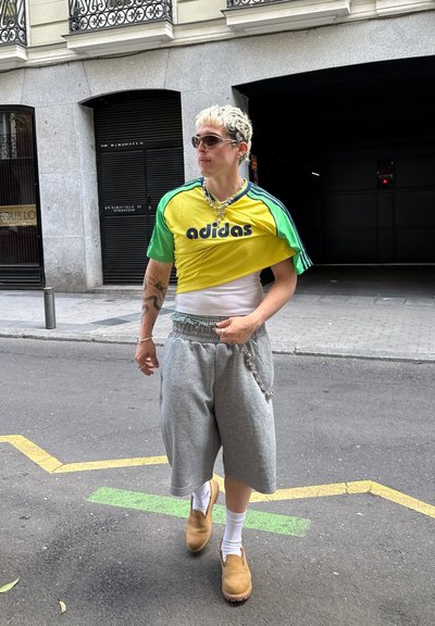 Jersey verde y amarillo de Adidas, pantalones cortos gris de gran tamaño, zuecos marrones, calcetines blancos y collar de cadena plateada, caminando por una calle con líneas amarillas.