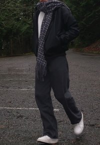 Veste bomber noire, chemise blanche, écharpe grise à carreaux, pantalon gris et baskets blanches ; debout sur une surface d'asphalte humide et sombre.