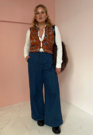 Chaleco de flores marrón sobre una camisa blanca, combinado con jeans de pierna ancha de cintura alta en azul y botas de plataforma negras. Bolsa de peluche marrón.