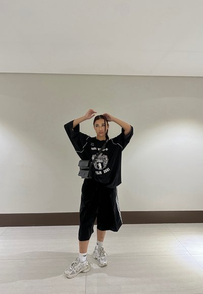 Camiseta negra de gran tamaño con diseño gráfico, combinada con pantalones cortos negros hasta la rodilla y zapatillas plateadas gruesas. Pequeño bolso negro bandolera.