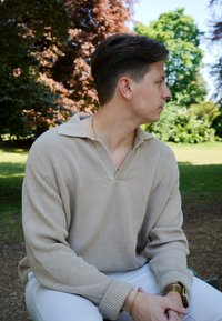 Pull en laine beige à col large, épaules tombantes et poignets côtelés, associé à un pantalon blanc et une montre-bracelet dorée.