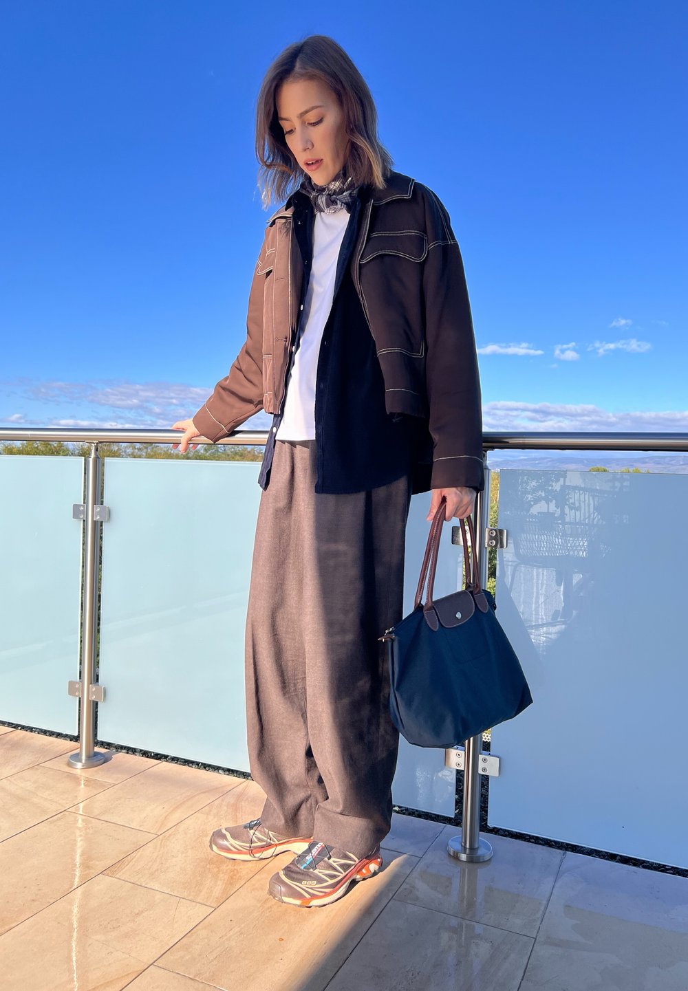 Καφέ κοντό τζάκετ πάνω από μπλε πουκάμισο, λευκό μπλουζάκι, γκρίζες φαρδιές παντελόνες κα�ι γκρίζα αθλητικά παπούτσια. Κρατάει μια σκούρα μπλε τσάντα με καφέ λεπτομέρειες.